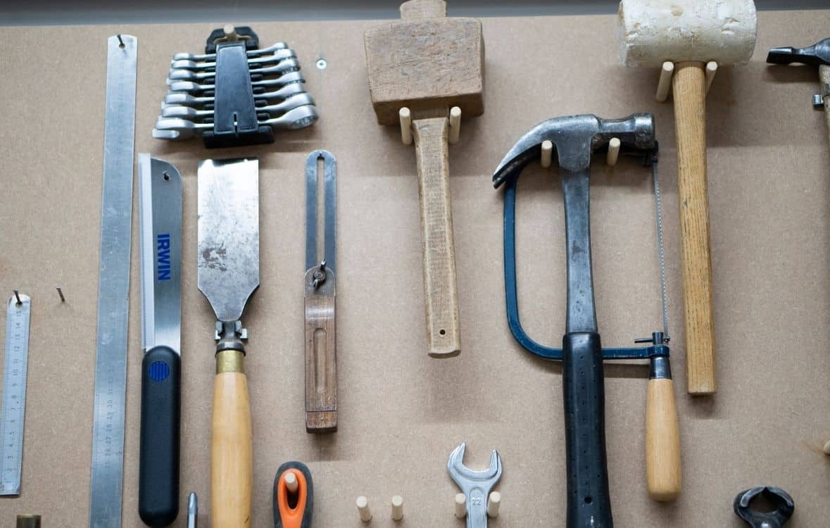Tools hanging on a wall, including hammers, a saw, and a wrench against a beige background.