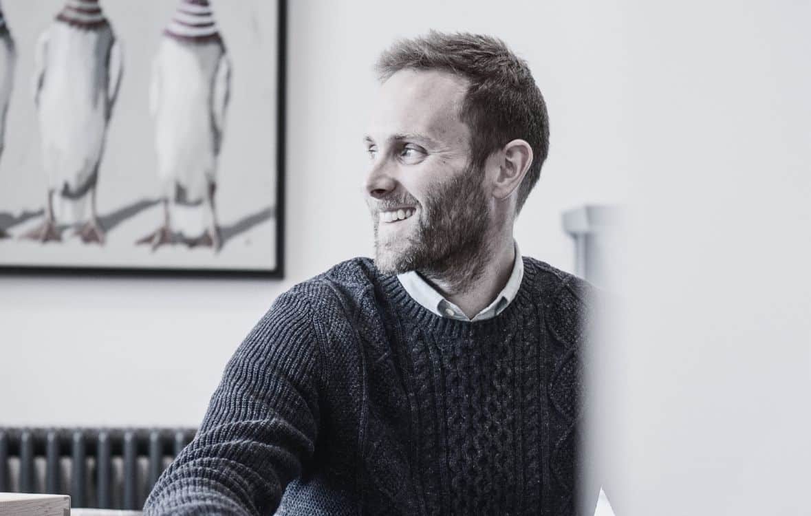 A man in a sweater smiles, sitting indoors. Three penguins in hats are depicted on the wall behind him.