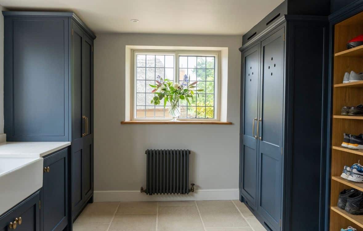 Laundry room with dark cabinets, a shoe shelf, and a window with a flower vase, above a radiator.