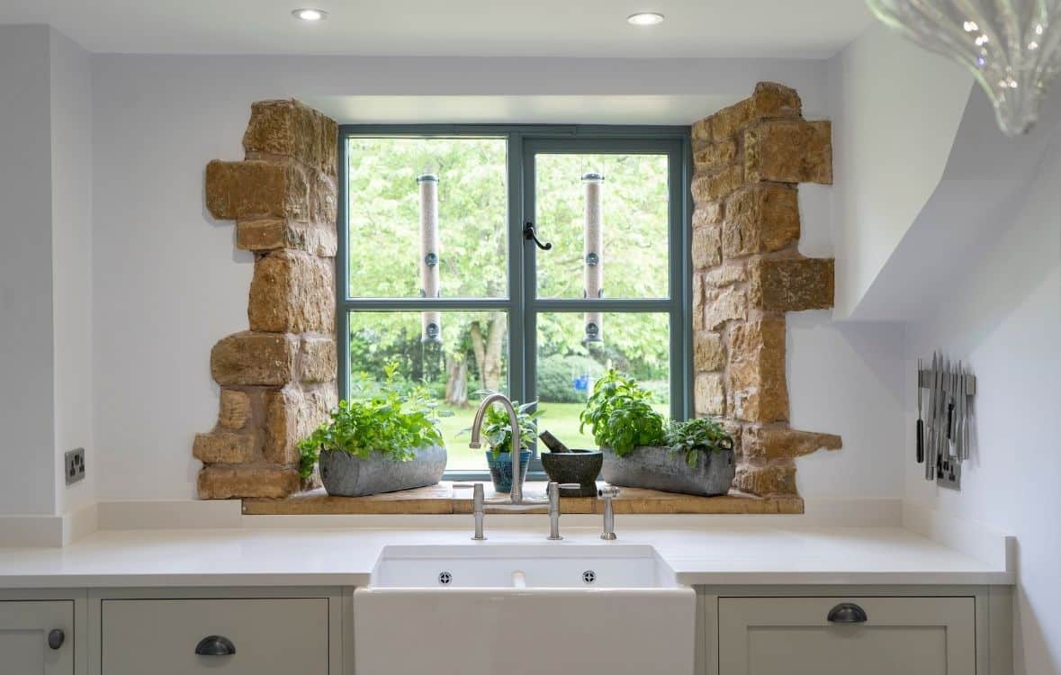 A rustic kitchen window with stone framing, plants on the sill, and bird feeders outside.