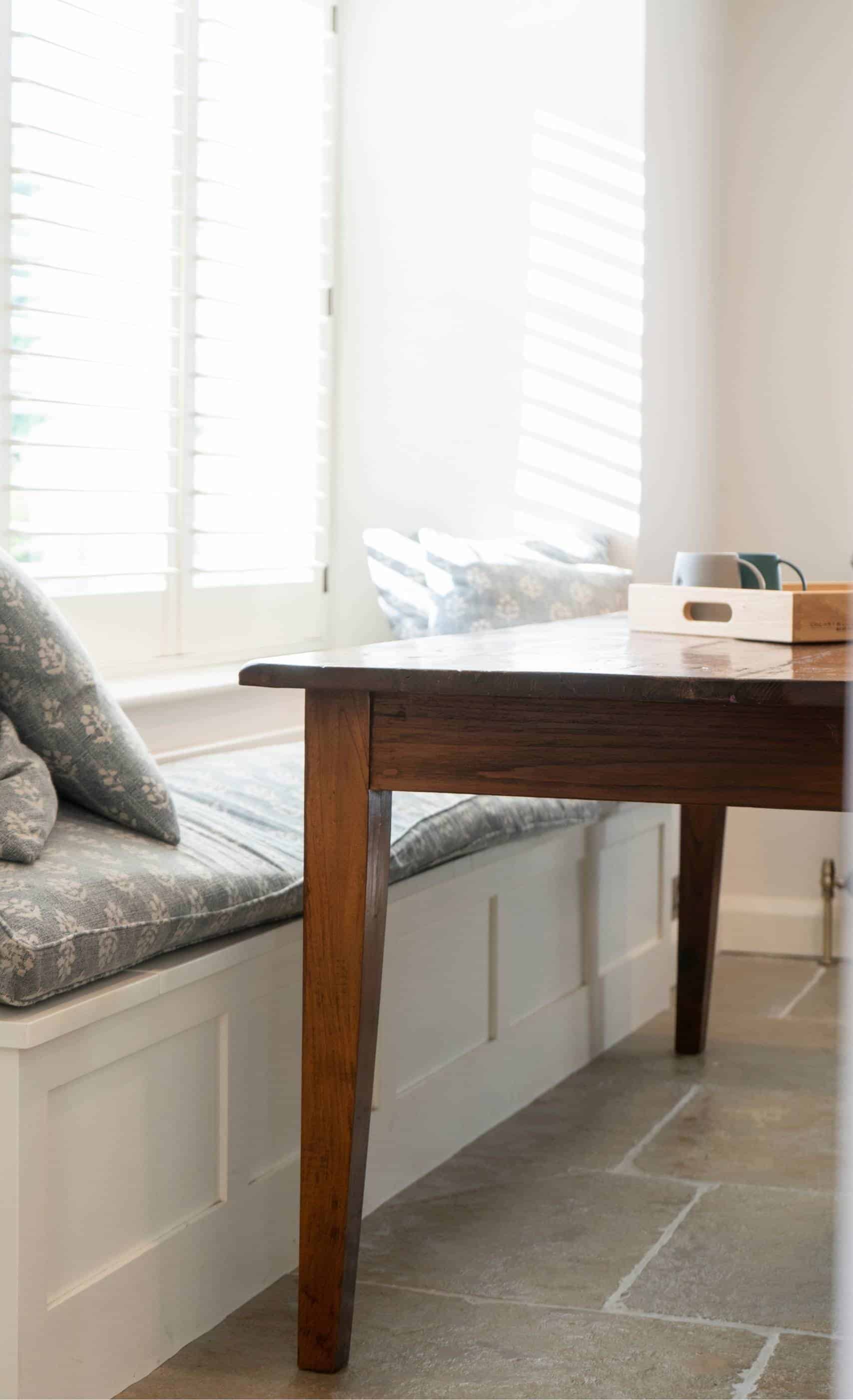 Cozy dining nook with a wooden table, bench, and cushions by a window with sunlight filtering through blinds.