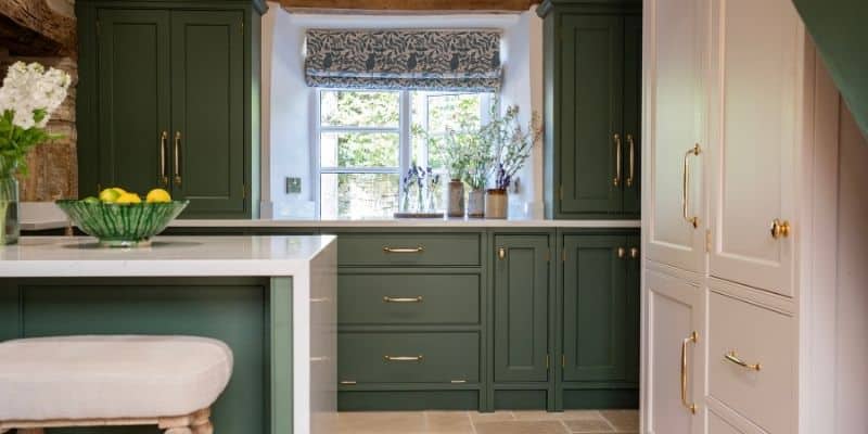 Cozy kitchen with green cabinets, white counters, and potted plants near a window with a patterned shade.