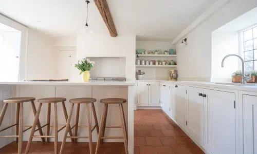 Bright kitchen with wooden stools, white cabinets, and terracotta floor. A vase with flowers is on the counter.