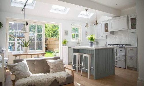 Bright kitchen with a central island, skylights, and plants, opening to a garden. Cozy sofa and dining area.