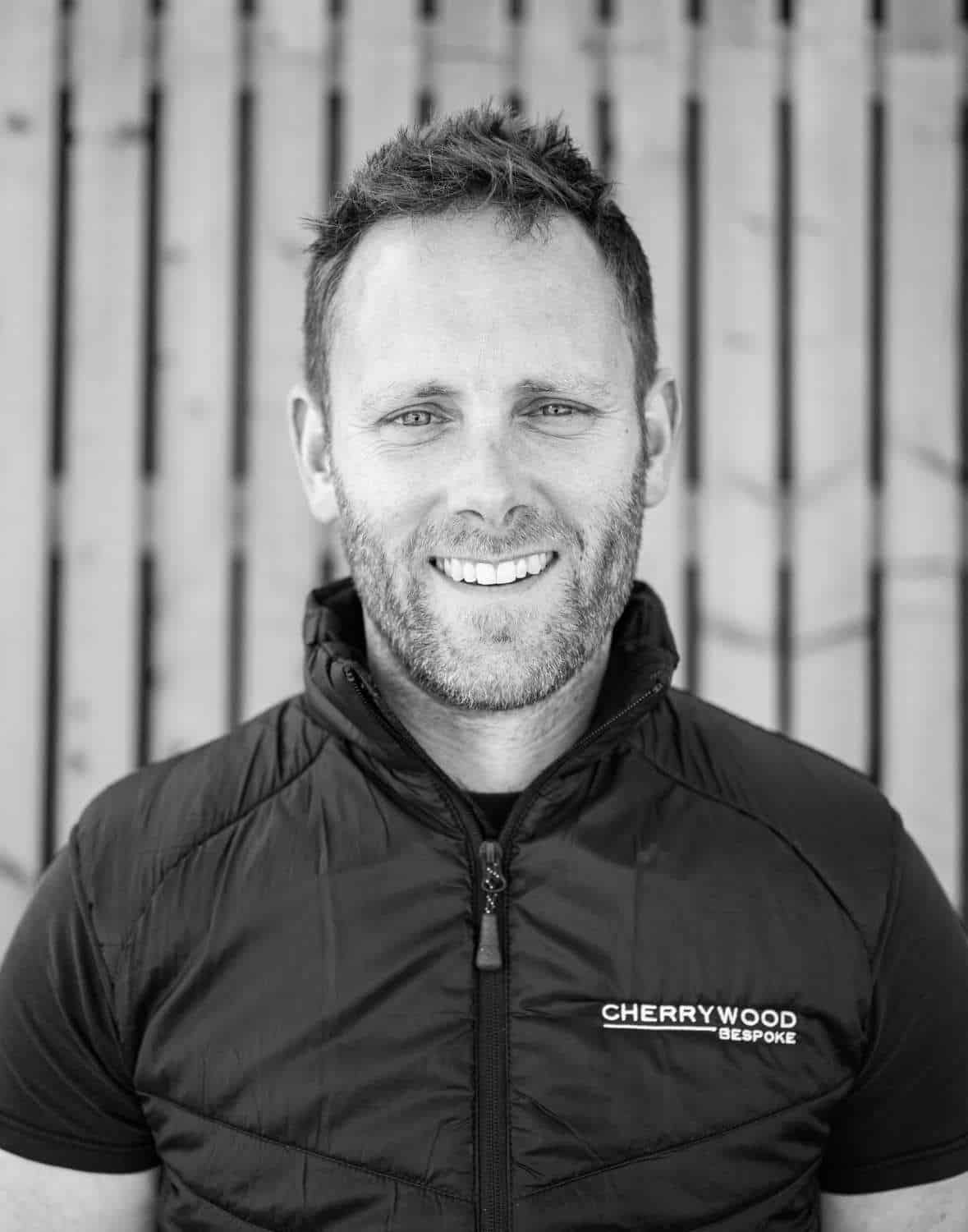 A man smiling, wearing a Cherrywood Bespoke jacket, standing in front of a wooden fence.