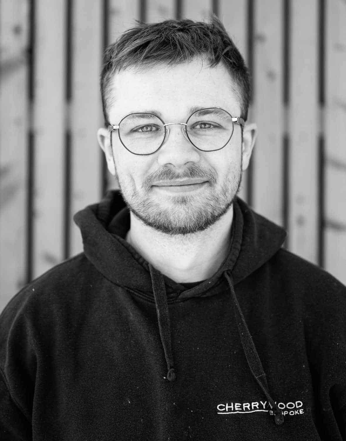 Man with glasses and beard wearing a hoodie, standing in front of a wooden background.
