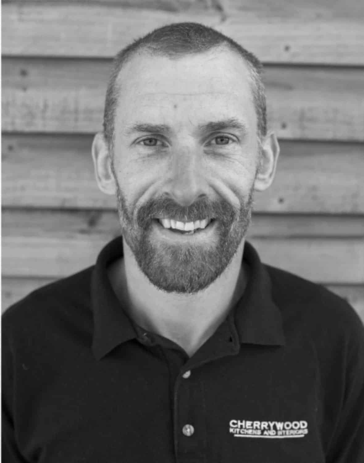 Man smiling in a black polo shirt against a wooden background.