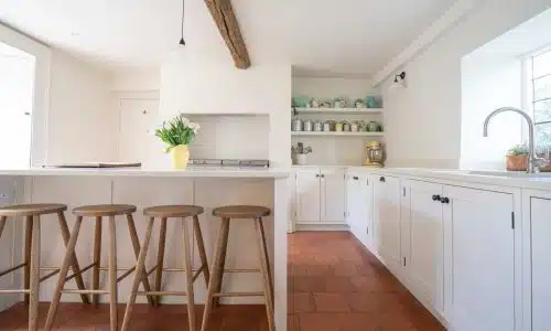 Bright kitchen with white cabinets, wooden stools, open shelves, and sunlight streaming through the windows.